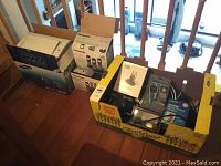 Wide shot of all items including Samsung printer box, Panasonic phone box, RCA clock radio box, Fellowes shredder box, and blood pressure monitor box placed on wooden floor.