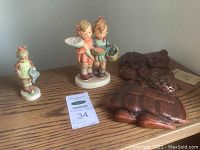 Photo showing three Hummel figurines and two dark brown carved animal figures on a wooden surface, includes auction tag.