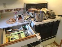 Wide view showing countertop with multiple pots and utensils on stove and drawer partially open with utensils inside.