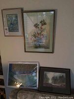 Overview photo of four framed artworks on a wall and leaning against a surface.