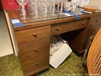 Front view of the wooden mid-century modern desk showing three drawers on the left and one drawer in the center with various glassware items on top covered with a protective glass panel.