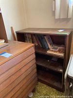 Wood bookshelf with three shelves containing assorted books and some papers, positioned against wall near window with vertical blinds.