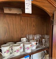 Wood cabinet shelf with six porcelain coffee cups with gold rim and floral designs on front, plus various clear aperitif and wine glasses behind