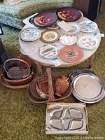 Top view of round table with multiple collectible plates featuring various artistic and scenic designs, metal platters, and several wicker baskets arranged around the table.