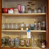 Glass cabinet shelves with an assortment of clear wine glasses, small clear tumblers, mugs including Santa Claus, and several collectible and decorative drinking glasses.