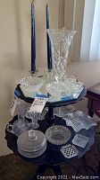 Clear vintage glassware on three-tier black stand including tall candles, vases, bowls, covered dish, and small decorative plates.