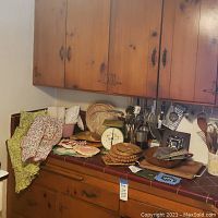 Wide view of kitchen countertop displaying various kitchen utensils, vintage food scale, baskets, trivets, aprons, and cloth items.