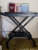 Front view of black rolling serving cart with two shelves, books and DVDs on top shelf, caster wheels beneath.