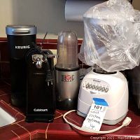 Photo showing four small kitchen appliances on countertop: Keurig coffee maker, Cuisinart kettle, Magic Bullet blender, Oster blender with jar covered in plastic bag.