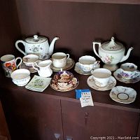 Full lot view showing two teapots, various teacups, saucers, small floral plates, and a creamer jug arranged on a wooden cabinet shelf.