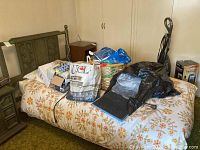 Full view of the twin bed frame with mattress covered in a floral bedspread and various bags on top.