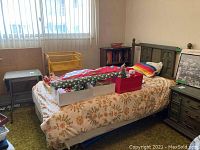 Twin brown metal bed frame with headboard and footboard along the left wall below a window with vertical blinds. Bed frame is unmade and has no mattress. Various objects on and near the bed are excluded.
