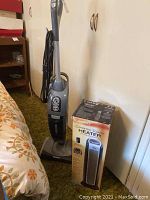 Front view of Oreck upright vacuum cleaner standing on carpeted floor beside bed and cabinet.
