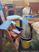 Photo showing two vintage wooden chairs with fabric seats, wooden stepping stool, metal fan in a yellow bucket, and basket with assorted contents.