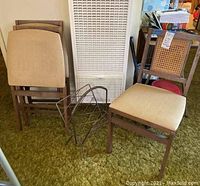 Image showing three folded vintage cane back chairs with beige vinyl seats stacked together beside a vintage metal magazine rack on green carpet.