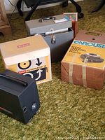 Wide shot showing three items on carpet: Kodak Automatic 8 Projector with box, Kodak Cavalcade Projector in case, and Kodak Carousel 850H Projector box.