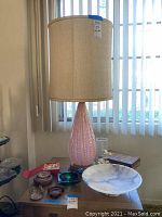 Vintage pink glass lamp with beige shade on table surrounded by various decorative items including marble bowl, wooden box, ashtrays, coral vase, shells, and coasters.