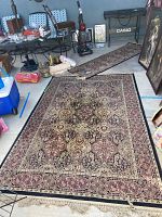 Photo showing the large area rug laid flat on the floor with intricate traditional pattern in red, beige, black and cream with fringe on one end.