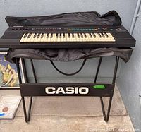 Casio keyboard on metal stand with brand name visible. Keyboard has black keys and several colored buttons, carrying case underneath.