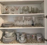 Full view of cabinet shelves showing assortment of Seagull China place settings, various coffee mugs, and multiple types of glassware including cocktail, wine, and steins.