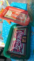 Red and green metal Coca Cola serving trays with vintage-style logos and graphics shown on a blue patterned table.