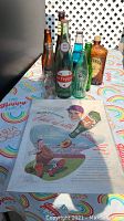 Photo showing collection of vintage glass soda bottles on a table with a colorful rainbow tablecloth and a vintage advertising poster partially under the bottles.