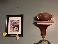 Framed Johnny Unitas print and Wilson NFL collectible football displayed on a wooden wall shelf with football player figurines.