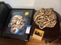 Photo showing lenticular Native American framed art, round plaster warrior battle cast, and small wooden plaque together on a dark surface.