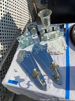 Photo of all items shown on a white tablecloth outdoors including two etched glass candle holders with metal mounts, mirrored glass clock, etched glass candle holders, glass vase, folded lace cloths, and a hanging mirror.