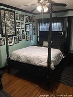 Full view of queen size black wooden four-poster bed with bedding on it, set up in a room with wooden floor and green walls with framed pictures.