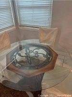 Round glass top dining table with octagonal wood and black metal scrollwork center and curved metal base on wood pedestal.