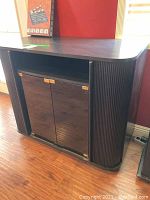 Front-side angle view of black stereo cabinet with glass doors, showing gold-tone hinges and handles, curved wood details on sides, and top surface condition.