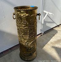 Full view of the brass tone embossed umbrella holder showing detailed scenes with buildings and a bridge, rope handles visible on the sides, standing on concrete floor outdoors.