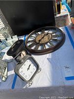 Three clocks arranged on a white tablecloth, including a large round wall clock and two smaller mantel clocks