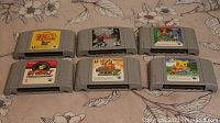 Photo showing six Japanese Nintendo 64 cartridges on floral fabric background, with visible labels including Donkey Kong 64, The Legend of Zelda, Pokemon Snap, and others.