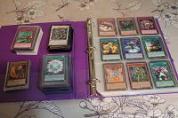 Open binder with rows of Yugioh cards visible in sleeves and stacks beside it on the left page.