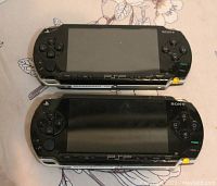 Photo showing two black Sony PSP consoles front view on floral patterned surface, displaying screen and buttons.