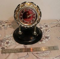 Side view of vintage USSR Majak clock showing red dial face with white numbers, gold accents, and faceted clear glass surround with star-like design. Mounted on black circular base with two chrome rods and balls.