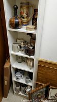 Front view of tall white shelving unit with shelves displaying beer steins, pottery mugs, wooden coasters on top shelf, and two framed wall decor pieces leaning at base.