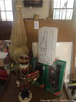 Full view of the Christmas decor lot showing the nutcracker holding a biplane, the tall gold mesh pointed tree decoration adorned with ribbons and ornaments, two boxed Dickens' Village figurines, and a boxed candle holder.