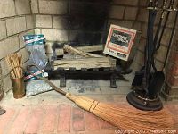Wide view of fireplace with cast iron grate, firewood, chimney brush box, broom, and fireplace tool set on stand