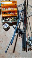 Photo shows an open yellow vintage fishing tackle box with compartments containing assorted fishing lures, hooks, sinkers, and other tackle. Several fishing rods with reels are placed to the right of the box, with a fishing net partially visible.