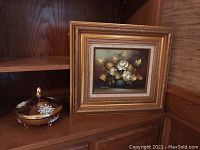 Full view of the framed flower painting and the decorative bowl on a wooden shelf.