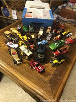 Photo showing a variety of approximately 22 die-cast toy cars arranged on a wooden table, including classic cars, race cars, and a distinctive yellow delivery van.