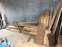 Wide view of a large pile of assorted wood pieces stacked on the floor against a concrete wall.