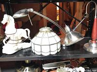 Wide view showing white ceramic horse lamp, Tiffany-style stained glass lamp shade, and adjustable metal desk lamp together on a black shelf.