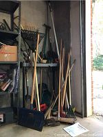 Photo showing a collection of yard tools including rakes, shovels, broom and a snow shovel leaned against a garage wall.