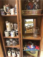 Full view of shelves showing multiple Oktoberfest beer steins in ceramic and stoneware, Margaritaville bar sign, kitchen plaque, and heraldic crests.