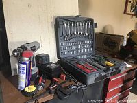 Photo showing cordless drill, battery, charger, WD-40 can, tape measure, flashlight, and mini tool kit opened with wrenches, sockets, screwdrivers, pliers, and clamp.