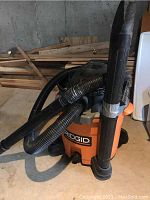Photo of Ridgid shop vacuum with black hose attached, extension wand taped on front, on concrete floor.